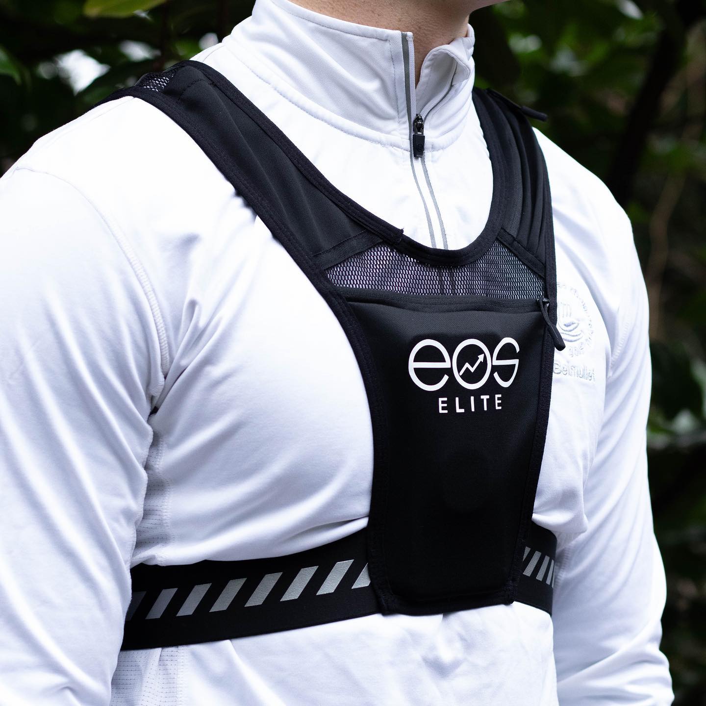A close up image of a male model wearing a black Eos Elite running vest over a white zipped running top is shown. The large front phone and strap pockets and elasticated band are clearly shown.