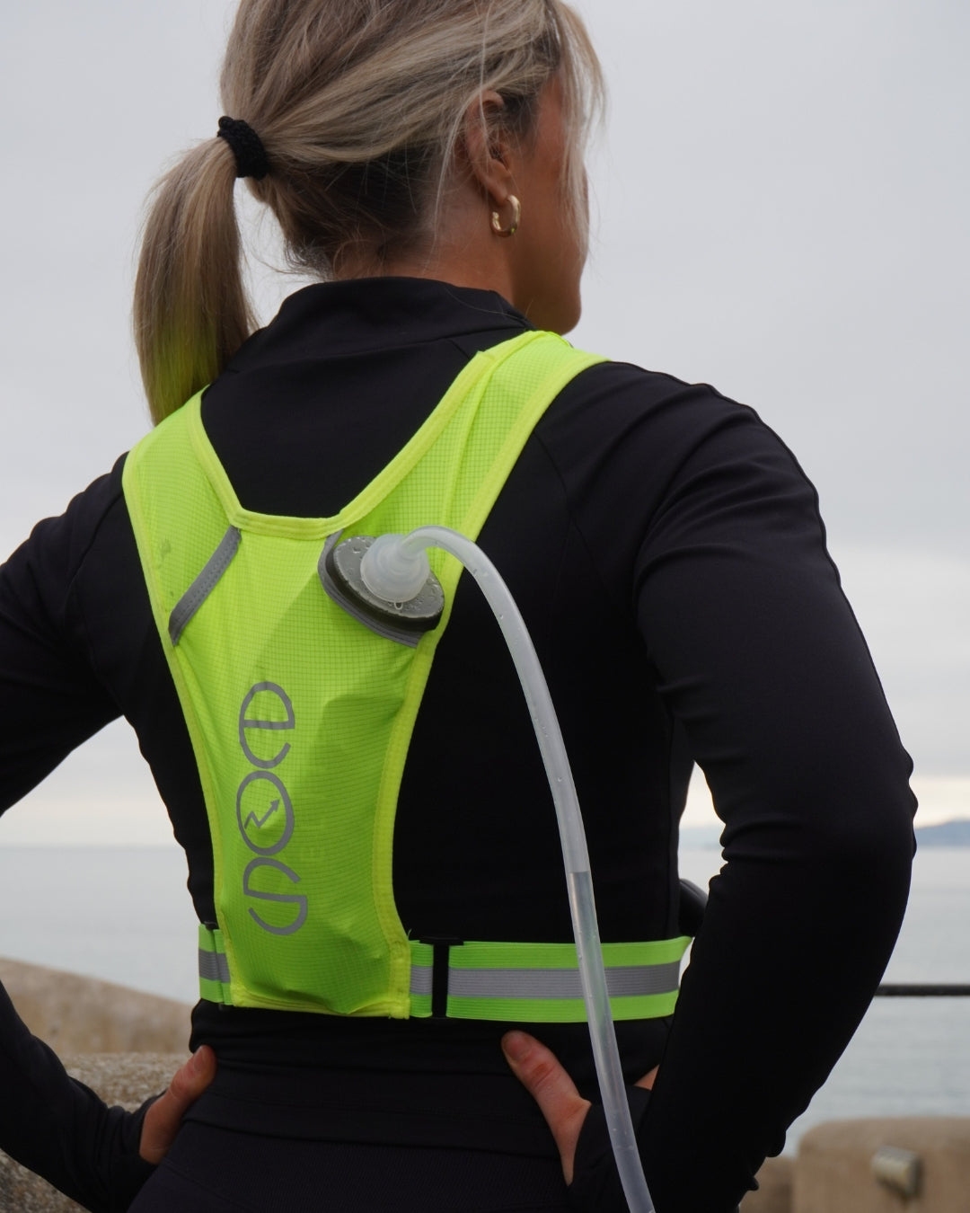 Eos Elite brand ambassador Anna Lalor is shown wearing the yellow Eos Elite IlluminLight running vest and water bladder. She looks out to sea with her back to the camera showing the reflective strips on the Eos Elite running vest.
