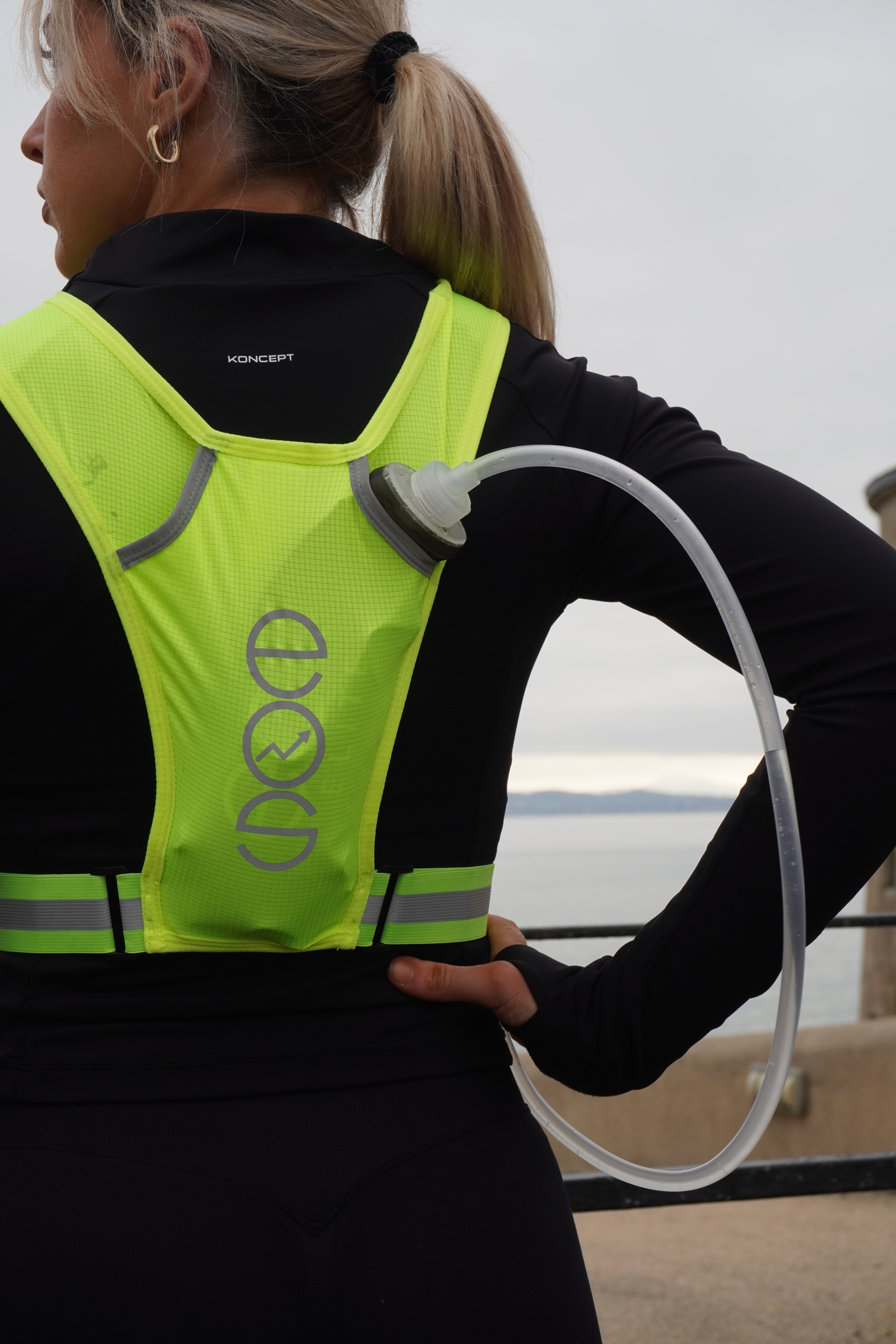 Eos Elite brand ambassador Anna Lalor is shown wearing the yellow Eos Elite IlluminLight running vest and water bladder. She looks out to sea with her back to the camera showing the reflective strips on the Eos Elite running vest.
