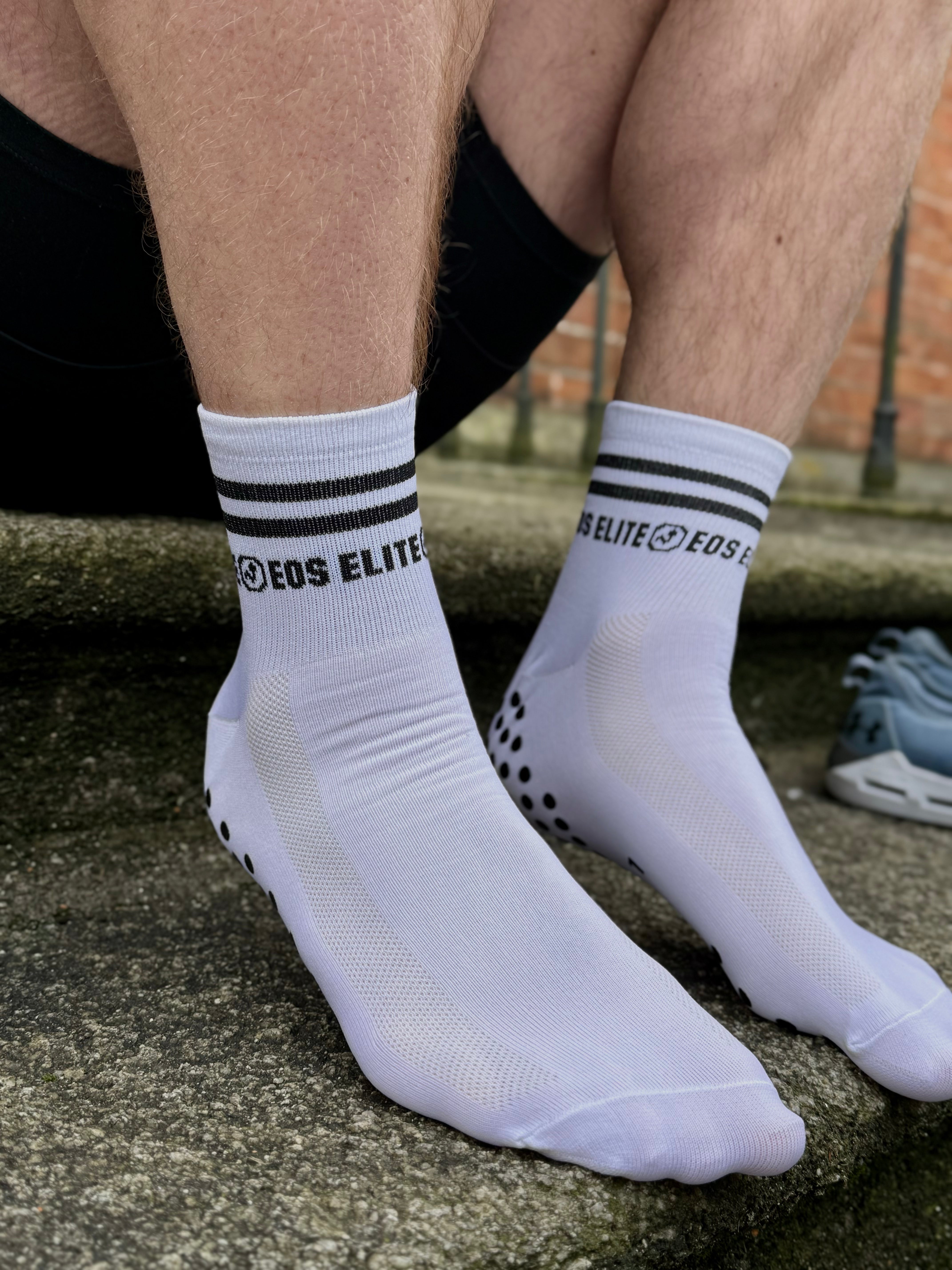 An arched foot on top of an outdoor step is shown in the image wearing a white Eos Elite Run Club grip sock. The silicon grip pads on the base of the sock used for ankle stability and comfor are clearly shown as is the brand's logo.