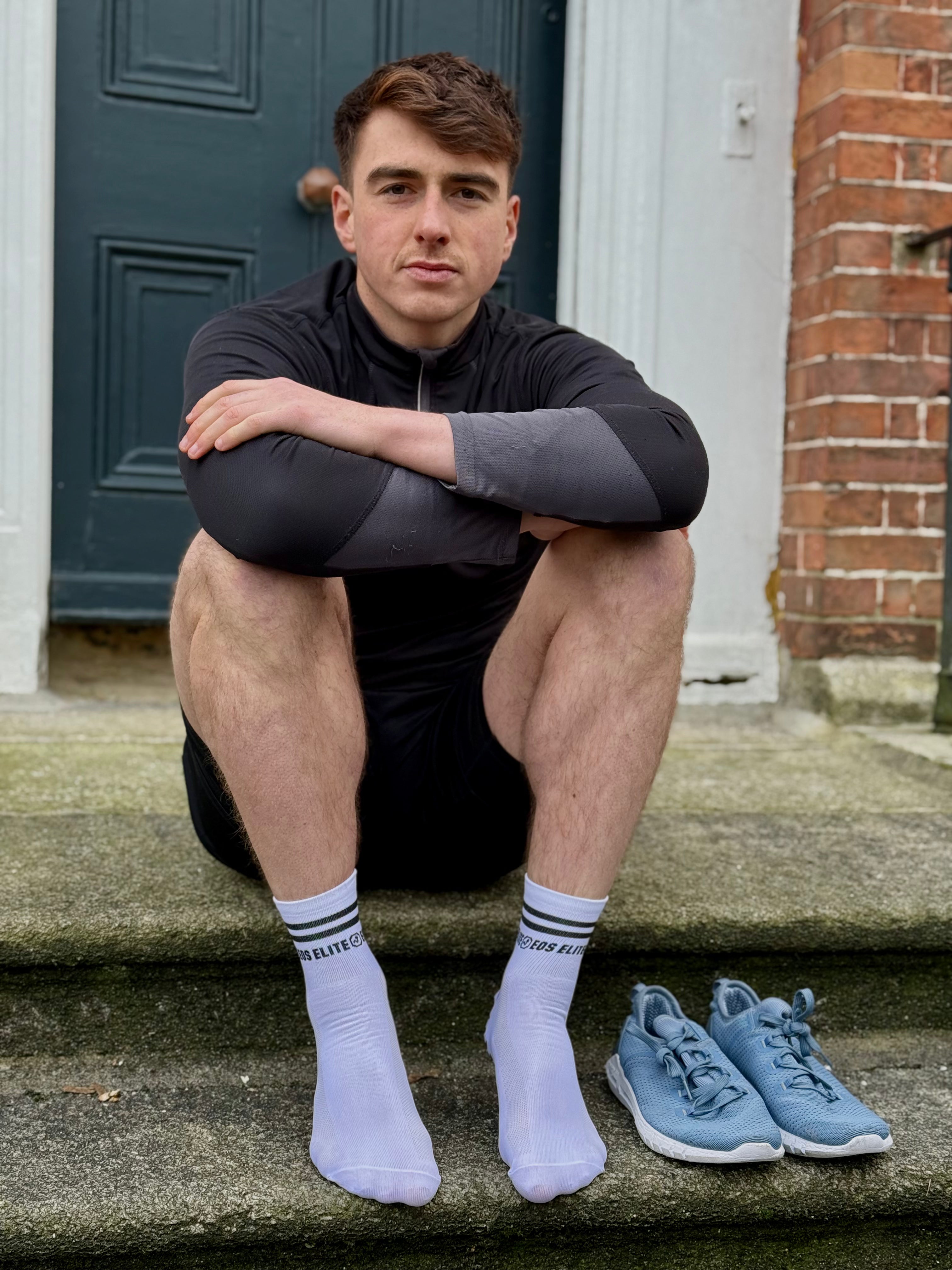Eos Elite Founder and CEO Lorcan Healy sits on a door step beside his pale blue training shoes. In focus are his white Eos Elite run club ankle length grip socks with silicon grip pads