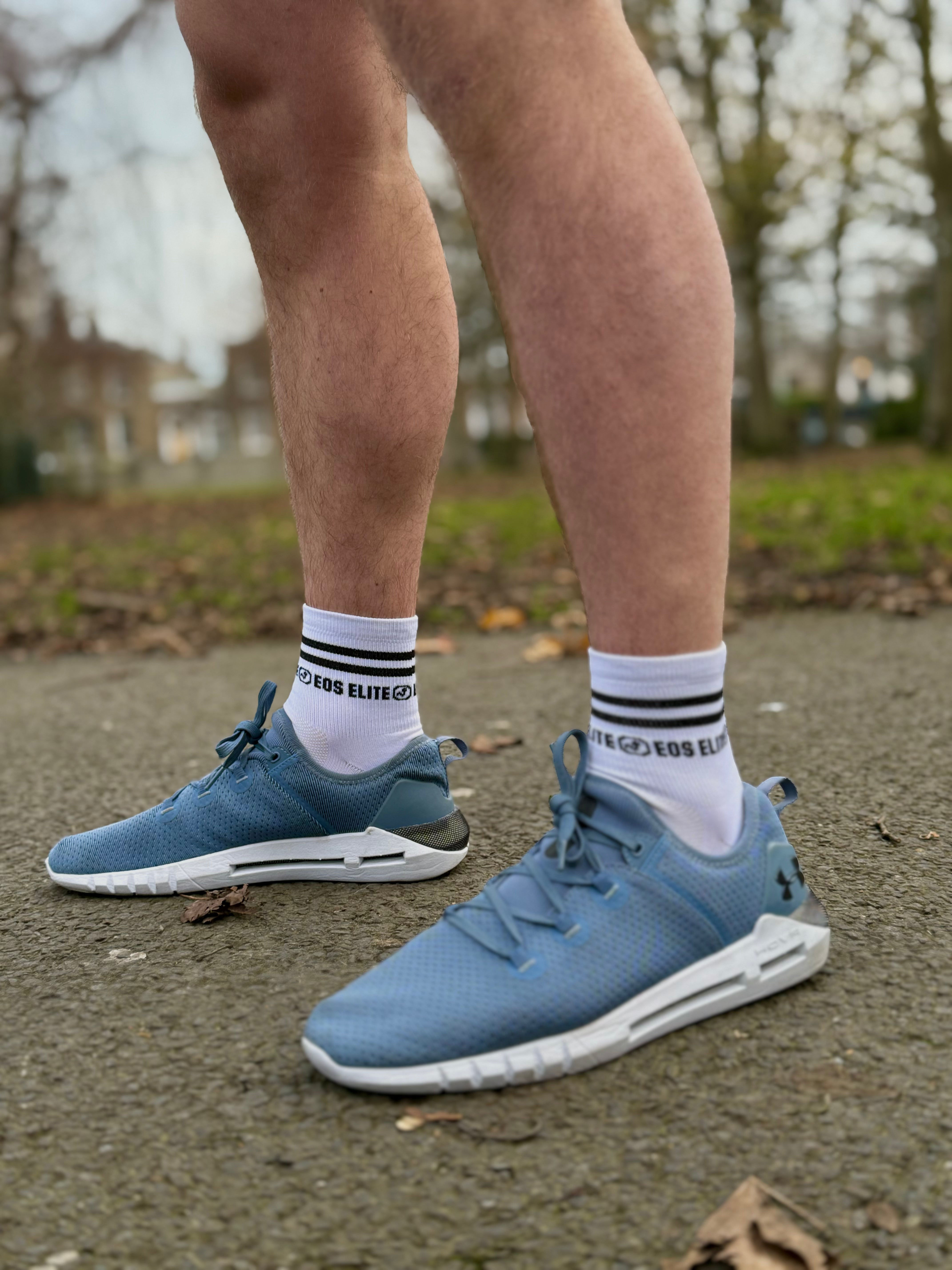 A close up image of runners legs in blue training shoes is shown. The focus of the image are the white Eos Elite run club grip socks worn.