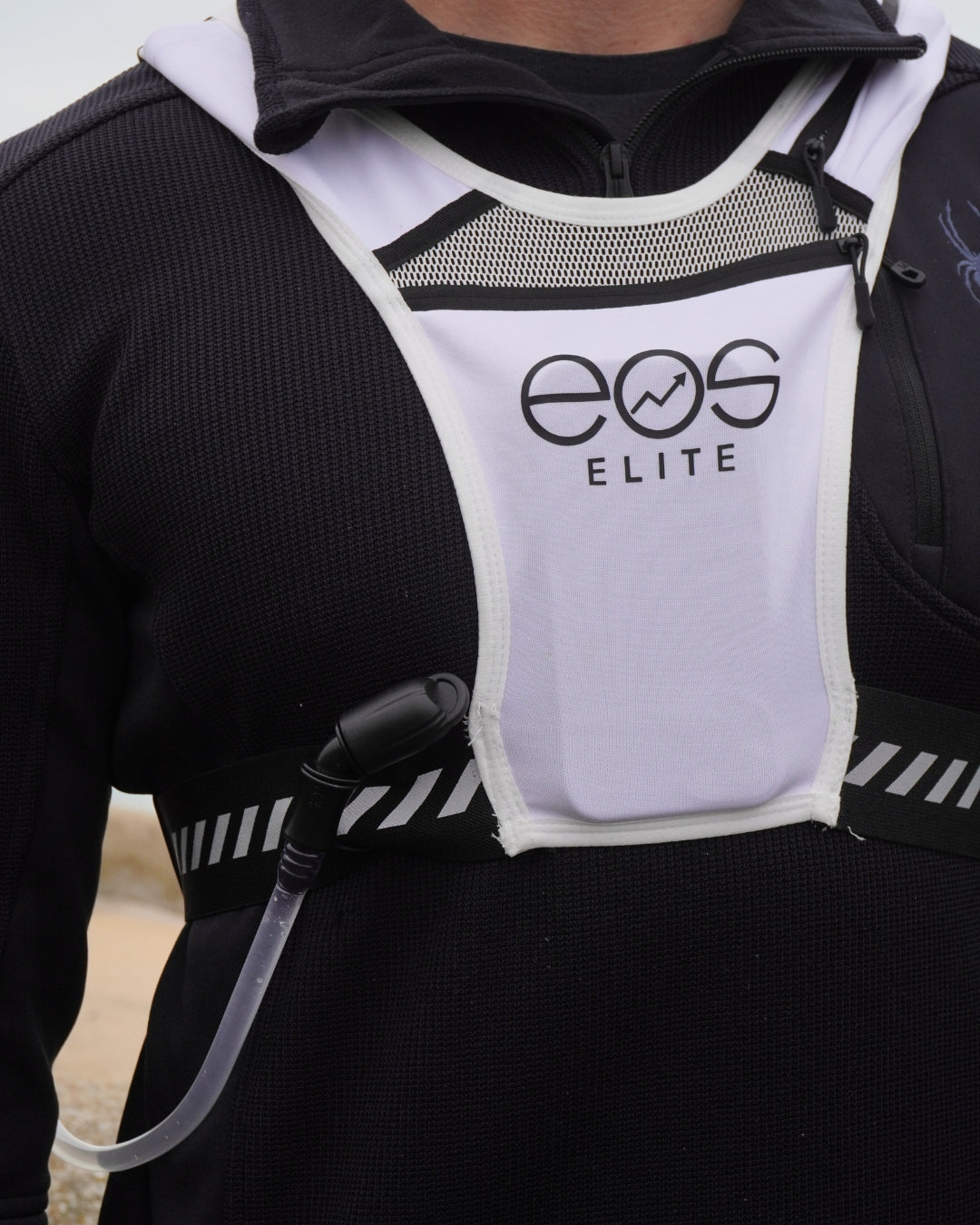 A male sportswear model wears a white Eos Elite running vest over a black long sleeved running top. The large front phone and strap pockets and adjustable straps are clearly visible.