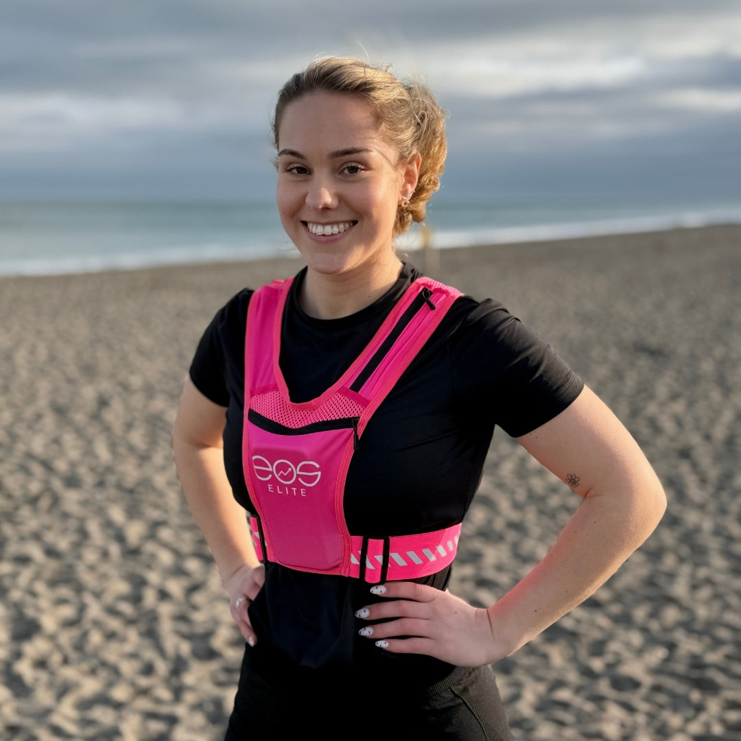 Eos Elite brand ambassador Molly Erin stands on a beach front of the ocean and smiles to the camera whilst wearing the pink Eos Elite  running vest. The front phone pocket, strap pocket and adjustable straps are clearly visible.