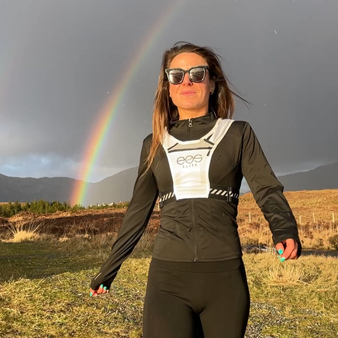 Eos Elite brand ambassador Sarah Murtag stands in front of grey clouds and a vivid rainbows  whilst wearing the white Eos Elite running vest. The front phone pocket, strap pocket and adjustable straps are clearly visible.