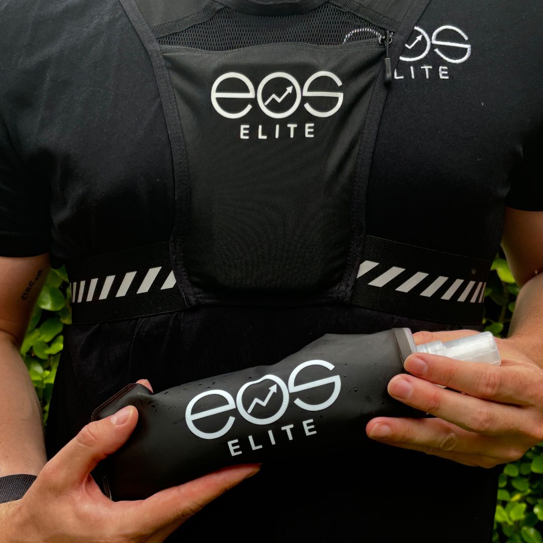 An athlete wearing a black Eos Elite running vest and holding a black Eos Elite silicon water bladder is shown. The adjustable straps, and large front phone pocket feature of the running vest are visible.