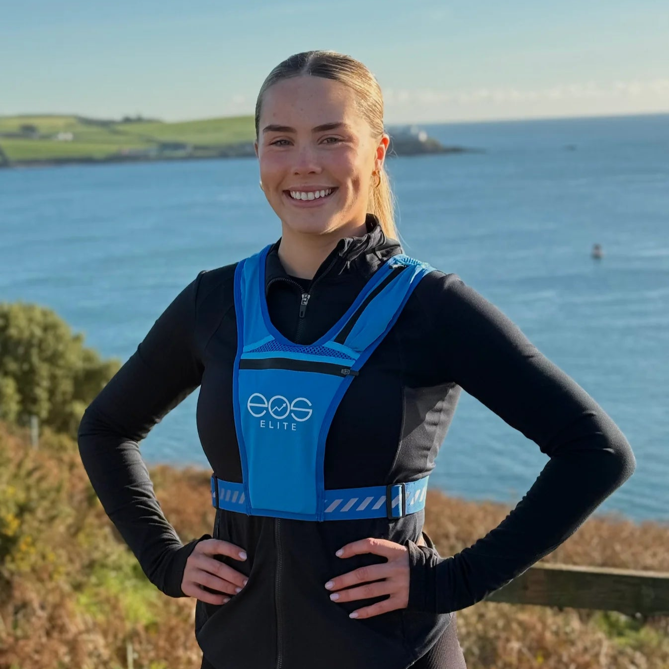 Eos Elite brand ambassador Erica Deasy stands in front of the ocean and smiles to the camera whilst wearing the blue Eos Elite waterproof RainGuard running vest. The front phone pocket, strap pocket and adjustable straps are clearly visible.
