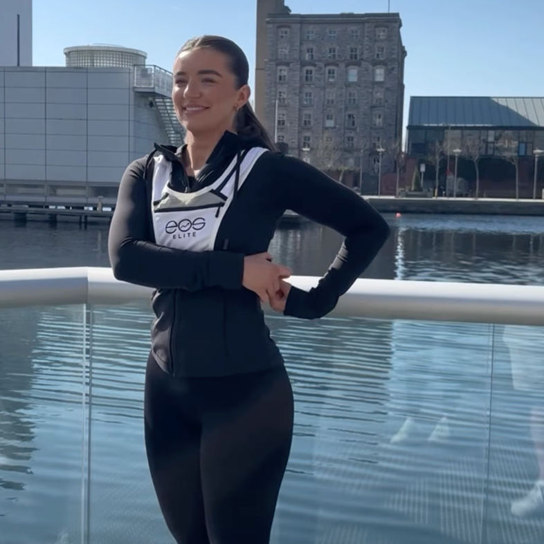 Eos Elite brand ambassador Jasmine Murray stands in front of the River Liffey and smiles whilst wearing the white Eos Elite running vest. The front phone pocket, strap pocket and adjustable straps are clearly visible.