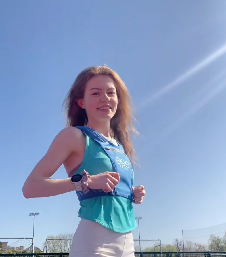 Eos Elite brand ambassador Elizabeth Gahan stands in front of blue skies on a running track smiles to the camera whilst wearing the blue Eos Elite waterproof RainGuard running vest. The front phone pocket, strap pocket and adjustable straps are clearly visible.