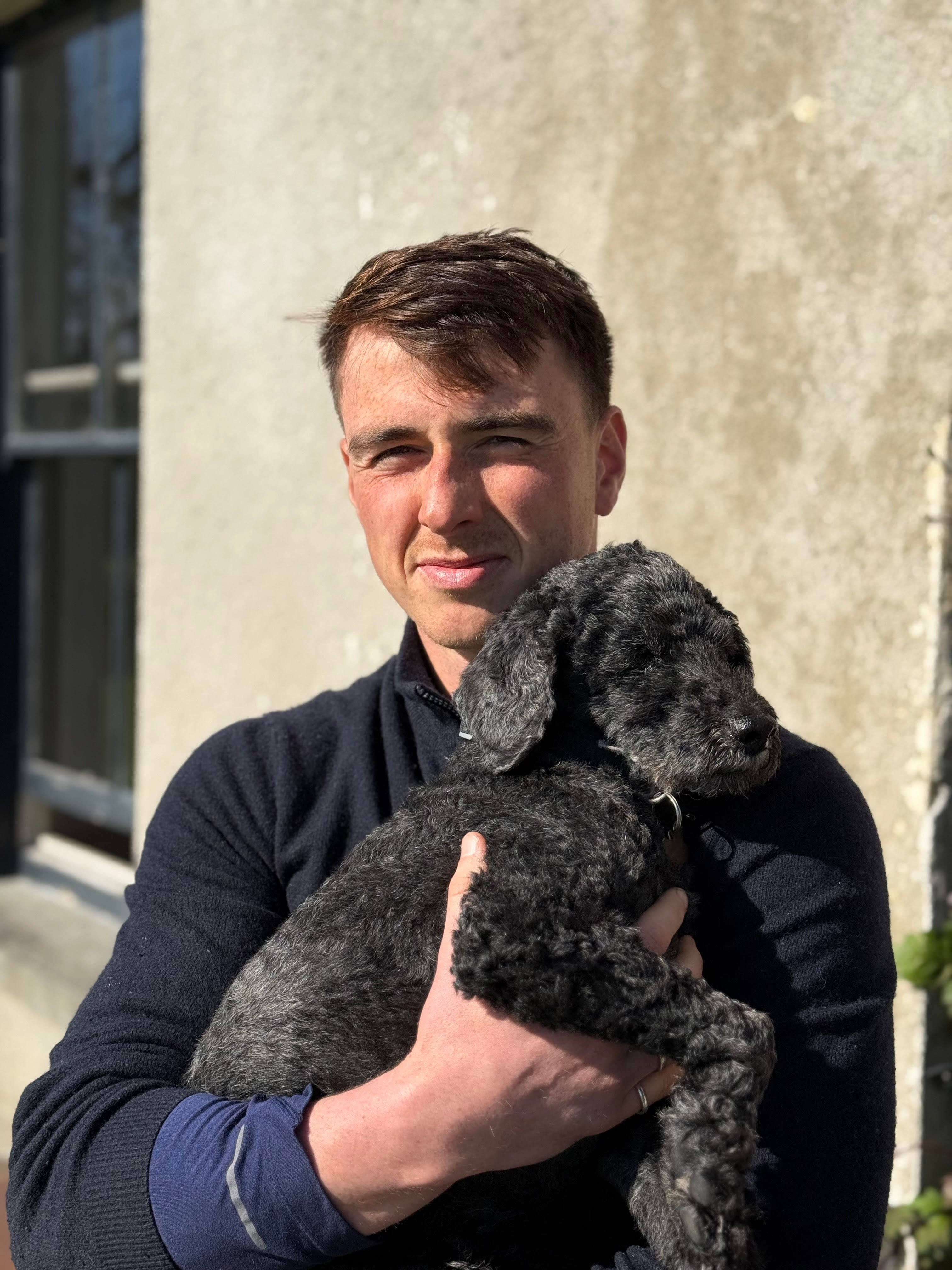 Eos Elite founder and CEO Lorcan Heany stands in front of a building holding a small, dark grey dog. A window behind them is slightly ajar and the sun is shining brightly.