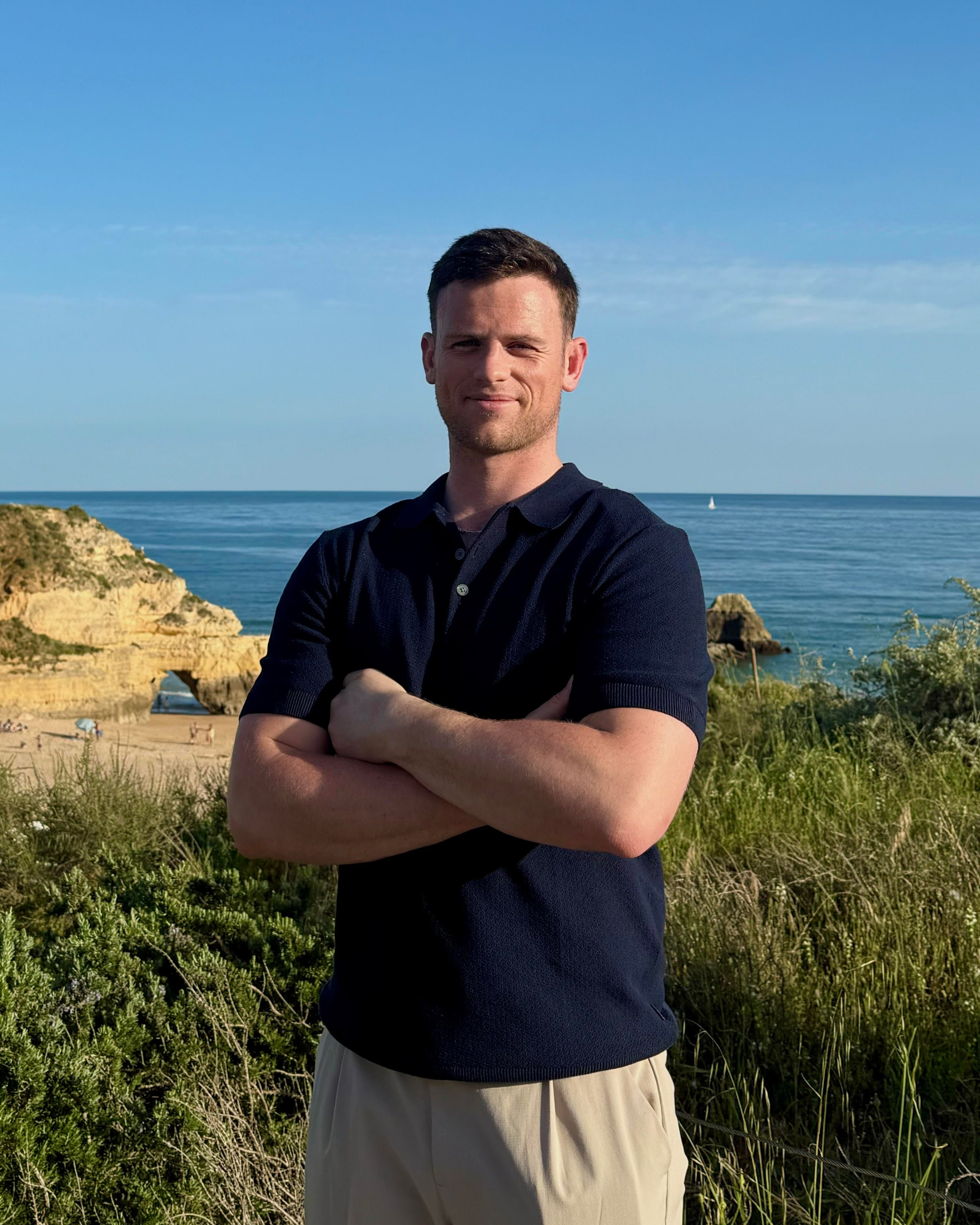 Simon Feeney, head of eCommerce for Eos Elite stands by the ocean and blue sky with folded arms smiling.